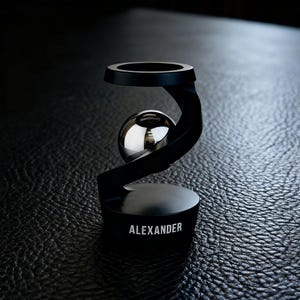 May include: A black metal desk accessory with a polished silver sphere held in a curved support. The base of the accessory has the name "ALEXANDER" in white lettering. The design is modern and minimalist.