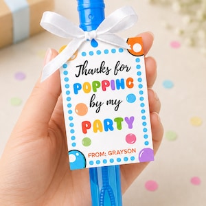 Op de afbeelding: Een blauwe bellenblaas met een wit label vastgebonden met een wit lint. Het label leest "Thanks for POPPING by my PARTY" in kleurrijke letters, met de naam "Grayson" onderaan. De bellenblaas wordt in een hand gehouden.