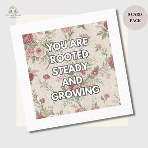 May include: A greeting card featuring a floral pattern with pink and red roses and the words "YOU ARE ROOTED STEADY AND GROWING". The card has a cream-colored border. The text is in a bold, black font.