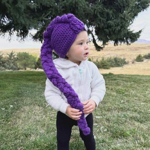May include: A vibrant purple crochet hat with a long, braided tail. The hat is a deep shade of purple and appears to be handmade. The hat is designed for a child and is a whimsical accessory.