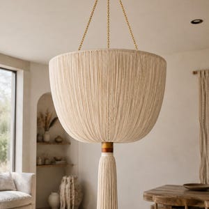 May include: A large, bowl-shaped pendant light fitting featuring a cream-coloured string-wrapped exterior. The fitting is suspended by three thin gold-toned chains from a matching circular ceiling canopy and includes a long, matching cream tassel.
