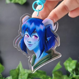 Puede incluir: Un llavero de acrílico transparente con una colorida ilustración de un personaje de piel azul con cabello azul oscuro, orejas puntiagudas y cuernos pequeños. El personaje tiene ojos morados y viste una prenda de color verde oscuro.