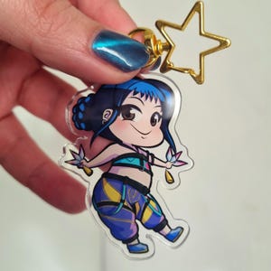 Può includere: Un portachiavi in acrilico trasparente con un personaggio dei cartoni animati con i capelli blu e un abito colorato. Il portachiavi ha un ciondolo a forma di stella dorata e una chiusura dorata. Il personaggio sorride e ha un design carino.