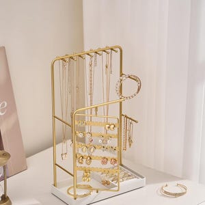 May include: Gold-coloured jewellery organiser with multiple tiers for necklaces, earrings, and bracelets. The organiser has a white base and a sleek, modern design. The word "LolaLet" is printed on the top right corner.