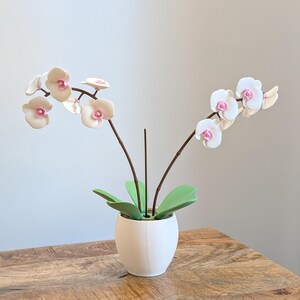 Może przedstawiać: Biały ceramiczny wazon zawiera sztuczną roślinę orchidei. Orchidea ma białe płatki z różowymi środkami, brązowe łodygi i zielone liście. Roślina stoi na drewnianym stole.
