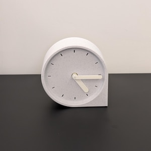 Könnte beinhalten: Eine moderne, minimalistische Schreibtischuhr in hellgrauer, gesprenkelter Ausführung. Die Uhr hat ein kreisförmiges Zifferblatt mit einfachen schwarzen Markierungen und weißen Zeigern. Das Gehäuse der Uhr hat ein einzigartiges, abgerundetes Design. Die Uhr ist ca. 15 cm breit.
