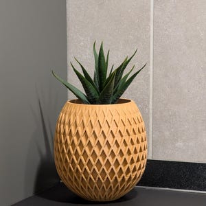 Op de afbeelding: Een decoratieve, lichtoranje ananasvormige plantenbak met een ruitpatroon. De plantenbak bevat een groene vetplant. De plantenbak staat op een zwart oppervlak, met een grijze muur op de achtergrond.