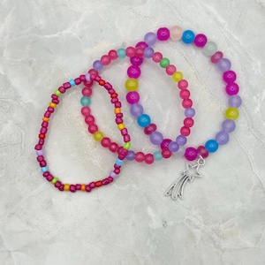 May include: Three colorful beaded bracelets. One bracelet is made of small red, yellow, and turquoise beads. The other two bracelets are made of larger pink, purple, blue, and yellow beads. One bracelet has a silver shooting star charm.