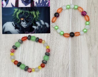 Puede incluir: Dos pulseras de cuentas con cuentas coloridas en tonos rojos, verdes, naranjas y transparentes. La imagen también incluye dos imágenes de personajes de anime. Las pulseras están sobre una superficie de vetas de madera clara.