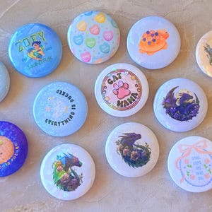Puede incluir: Una colección de botones redondos y coloridos con varios diseños. Algunos botones presentan ilustraciones de un pimiento, una tortuga, un dragón y una pata de gato. Otros botones tienen texto como "Zoey", "Cat Mama" y "Everything is ducked".