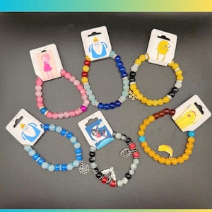 Könnte beinhalten: Auswahl an Armbändern mit Perlen und Cartoon-Charms. Jedes Armband hat ein einzigartiges Farbschema, darunter Rosa, Blau, Gelb und Schwarz, mit dekorativen Perlen und Charms. Die Armbänder werden auf einer dunklen Oberfläche präsentiert.