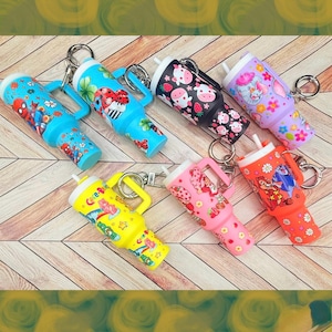 May include: A selection of miniature tumbler keyrings in various colours. Designs include cartoon characters and strawberries. Each tumbler has a handle and a straw, ideal for carrying small amounts of liquid. The keyrings are arranged on a wooden surface.