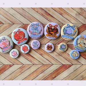 May include: A collection of colorful book club themed pinback buttons. The buttons feature illustrations and text such as "Romance", "Spicy", "Fantasy", "True Crime", "Magical", and "Mystery". The buttons are arranged on a wooden surface.