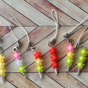 Könnte beinhalten: Eine Sammlung bunter sternförmiger Anhänger mit silberfarbenen Metallverschlüssen und weißen Kordeln. Die Anhänger haben verschiedene Farben, darunter Gelb, Rosa, Rot und Grün. Der Hintergrund ist eine hellbraune Holzoberfläche.