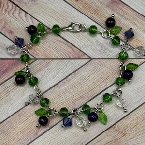 Könnte beinhalten: Ein filigranes Armband mit einer silbernen Kette, verziert mit grünen, klaren und dunkelvioletten Perlen. Das Armband enthält kleine grüne Blattanhänger und facettierte blaue und klare Kristallakzente. Der Verschluss ist silberfarben.