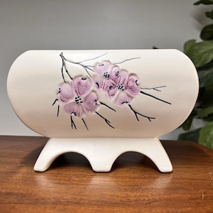 Può includere: Vaso in ceramica color crema con un motivo floreale in rilievo. Il vaso presenta tre fiori rosa con steli neri ed è sostenuto da tre piedi curvi. Il vaso ha una forma oblunga arrotondata ed è adatto per esporre fiori o come elemento decorativo.
