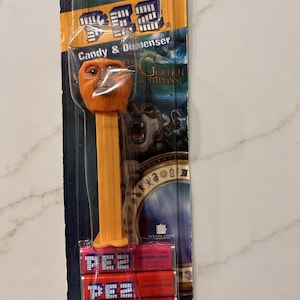 May include: An orange PEZ candy dispenser featuring a character from "The Golden Compass" movie, packaged with three rolls of raspberry, strawberry, and lemon candy. The packaging includes the PEZ logo and website address.