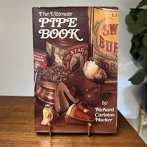 May include: The cover of "The Ultimate Pipe Book" by Richard Carleton Hacker, featuring a still life of pipes, tobacco, and related accessories. The book's title is in large, ornate lettering. A vintage tobacco tin is visible.