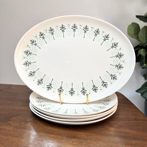 May include: A set of white ceramic plates with a decorative pattern of black and light green leaves and swirls. The plates are oval and round, with a gold-coloured stand holding the top plate.