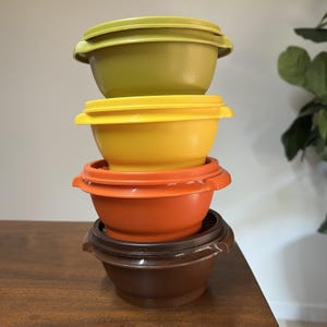 Peut inclure: Une pile de quatre bols en plastique colorés avec des couvercles assortis. Les bols sont dans des tons de vert, de jaune, d'orange et de marron. Les bols sont empilés les uns sur les autres.