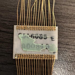 May include: A rectangular electronic component with a light gray surface featuring black printed text partially obscured by a light green adhesive patch. Thin gold-colored wire leads extend from the top and bottom edges of the component.