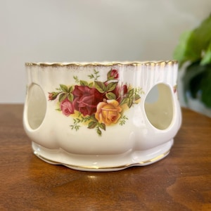 May include: A cream-colored ceramic tea light holder with a scalloped rim and gold trim. The exterior features a floral pattern of red, pink, and yellow roses with green leaves. The cylindrical body has three large circular cutouts for ventilation.