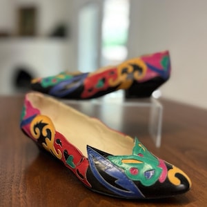 May include: A pair of low-heeled shoes featuring a black base with vibrant, multi-colored leather overlays in shades of red, yellow, green, and blue. The shoes have a pointed toe, a scalloped topline, and a low, solid black block heel.