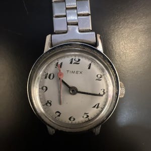 Könnte beinhalten: Eine silberfarbene analoge Armbanduhr mit rundem weißem Zifferblatt, schwarzen arabischen Ziffern und dem in Schwarz gedruckten Markennamen Timex. Die Uhr verfügt über ein schmales silbernes Metallgliederarmband, einen schwarzen Stundenzeiger, einen schwarzen Minutenzeiger und einen rosafarbenen Sekundenzeiger.