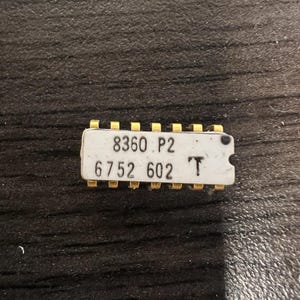 Puede incluir: Un chip de circuito integrado de cerámica blanca con pines metálicos dorados a lo largo de los bordes superior e inferior. La superficie superior presenta texto impreso en negro que dice 8360 P2 y 6752 602, junto con un pequeño punto negro y un símbolo T.