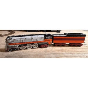Op de afbeelding: Een vintage speelgoedtreinset met een zilveren, oranje en zwarte locomotief en een bijpassende oranje en zwarte wagon. De trein staat op een houten oppervlak.