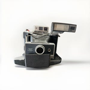 May include: A vintage Polaroid Land Camera 360 Electronic Flash camera. The camera is black and silver with a prominent lens and flash unit. The camera has the words "Land Camera 360 Electronic Flash" printed on the front.