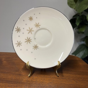 Peut inclure: Soucoupe crème avec un bord argenté et un motif d'éclats d'étoiles dorés. La soucoupe est présentée sur un présentoir en métal doré. Une petite indentation ronde est au centre.