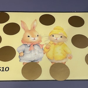 May include: A light yellow scratch-off card featuring two cartoon characters: a rabbit and a mouse. The rabbit wears a white outfit and a pink scarf, while the mouse wears a yellow jacket and hat. The card has brown scratch-off circles and an image of a golden egg with the text "=$10".