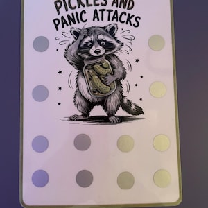 May include: A rectangular card with a green border features a cartoon raccoon clutching a jar of pickles. The text "PICKLES AND PANIC ATTACKS" is above the raccoon. The card has several gray and light green circles.