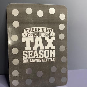 May include: A rectangular, gray sign with silver polka dots around the edges. The sign features the text "THERE'S NO CRYING DURING TAX SEASON (OK, MAYBE A LITTLE)" in white, bold font.