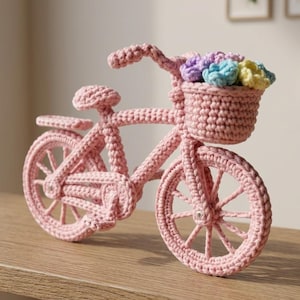 Może przedstawiać: Różowy szydełkowy rower z koszem pełnym kolorowych kwiatów. Ręcznie robiony rower posiada szczegółowe szprychy, siodełko, kierownicę i kosz. Przedmiot dekoracyjny jest eksponowany na drewnianej półce.