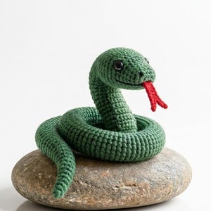 Op de afbeelding: Een handgemaakte groene gehaakte slang met een rode tong, zittend op een grijze steen. De slang heeft zwarte ogen en is opgerold. De achtergrond is wit.