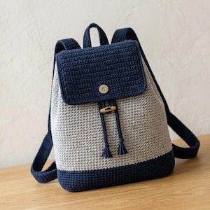 May include: A two-tone crochet backpack featuring a light gray main body and a dark navy blue base, top flap, and shoulder straps. The flap secures with a silver-toned metal snap, and the main compartment closes with a navy drawstring and wooden toggle.