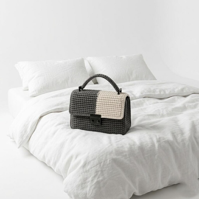 May include: A handmade crochet handbag in a two-tone design. The bag features a dark gray body with a cream-colored flap. It has a matching handle and a black clasp. The bag is sitting on a white bed.