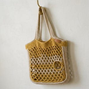 Könnte beinhalten: Eine Häkeltasche in Senfgelb und Beige. Die Tasche verfügt über ein rautenförmiges Netzmuster in der Mitte, feste Häkelränder, zwei Schulterriemen und ein kleines, rechteckiges Lederetikett an der oberen rechten Seite der Tasche.