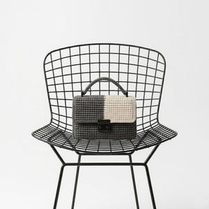 May include: A two-toned crochet handbag in gray and cream, with a black clasp and handle, rests on a black metal grid chair. The bag's design is split vertically, with one side gray and the other cream.