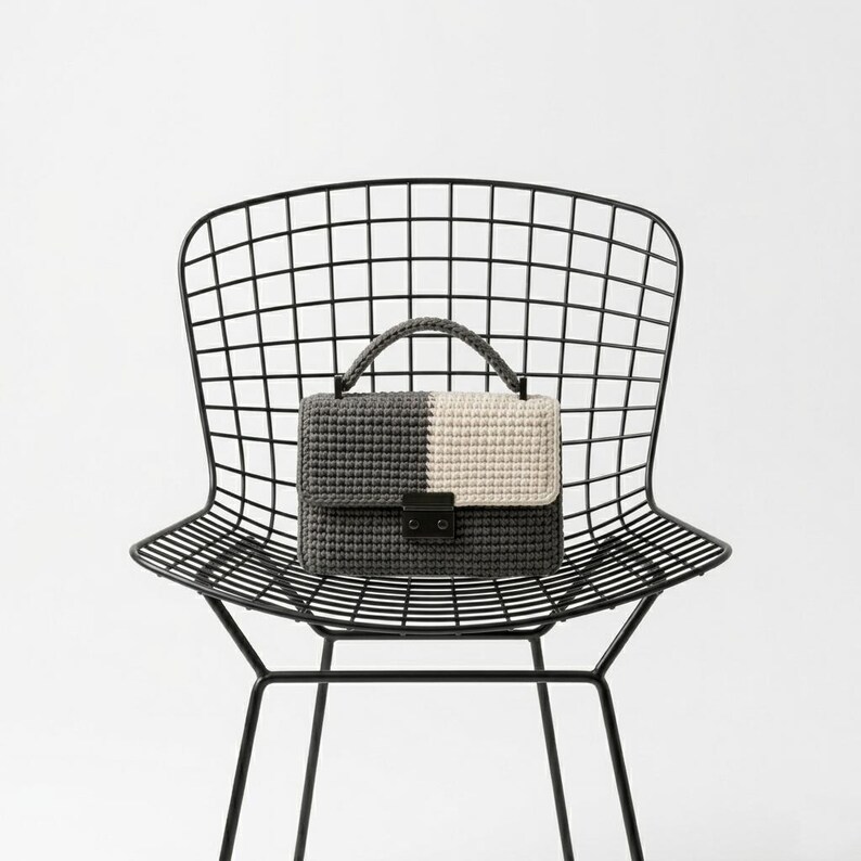 May include: A two-toned crochet handbag in gray and cream, with a black clasp and handle, rests on a black metal grid chair. The bag's design is split vertically, with one side gray and the other cream.