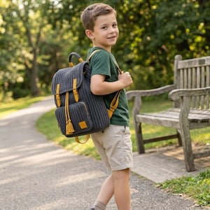 May include: A dark grey knitted backpack featuring a flap top with yellow leather-like straps, a front pocket with a yellow tassel drawstring, and adjustable yellow shoulder straps, worn by a child in a green t-shirt and beige shorts.