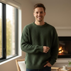 May include: A person wearing a dark green waffle-knit sweater. The sweater has a crew neck and long sleeves. The person is smiling and standing in front of a window and a fireplace.