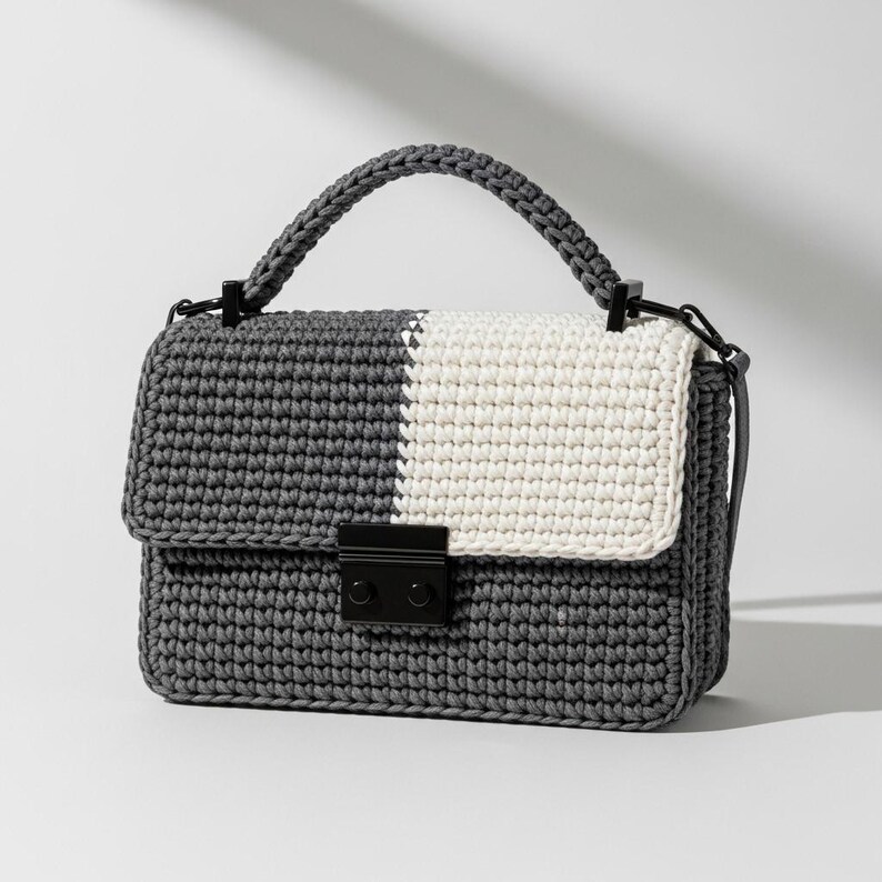 May include: A gray and white crochet handbag with a structured design. The bag features a flap closure with a black clasp and a matching handle. The bag's body is gray, with a white panel on the flap.