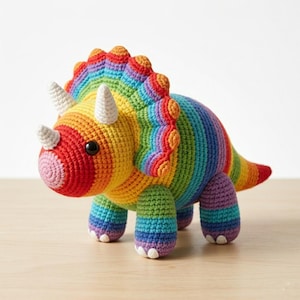 Op de afbeelding: Een gehaakte triceratops-dinosaurus in regenboogkleuren. Het handgemaakte speelgoed heeft een rode snuit, witte hoorns en een kleurrijke kraag. De dinosaurus heeft een afgerond lichaam en vier stevige poten.