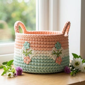May include: A handmade crochet basket with a peach, white, and mint green color scheme. The basket features two handles and is decorated with embroidered flowers. The basket is sitting on a wooden surface.