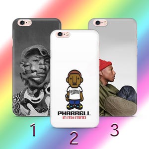 Puede incluir: Tres fundas para teléfono con diferentes diseños. Una funda es en blanco y negro con un retrato. Otra es blanca con una figura de dibujos animados y el texto "Pharrell In My Mind". La tercera funda es gris con una persona con un gorro rojo.