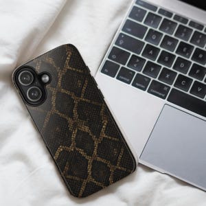 May include: A smartphone case with a snakeskin pattern in shades of brown and gold rests on a white surface next to a laptop. The case has a dual-lens camera and a sleek design, offering both style and protection.