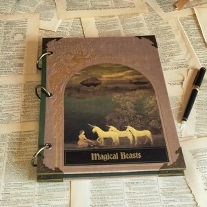 May include: A journal with a cover featuring a painting of unicorns and the text "Magical Beasts." The journal is open on a surface covered with old book pages, with a pen resting nearby. A collection of antique books is in the background.